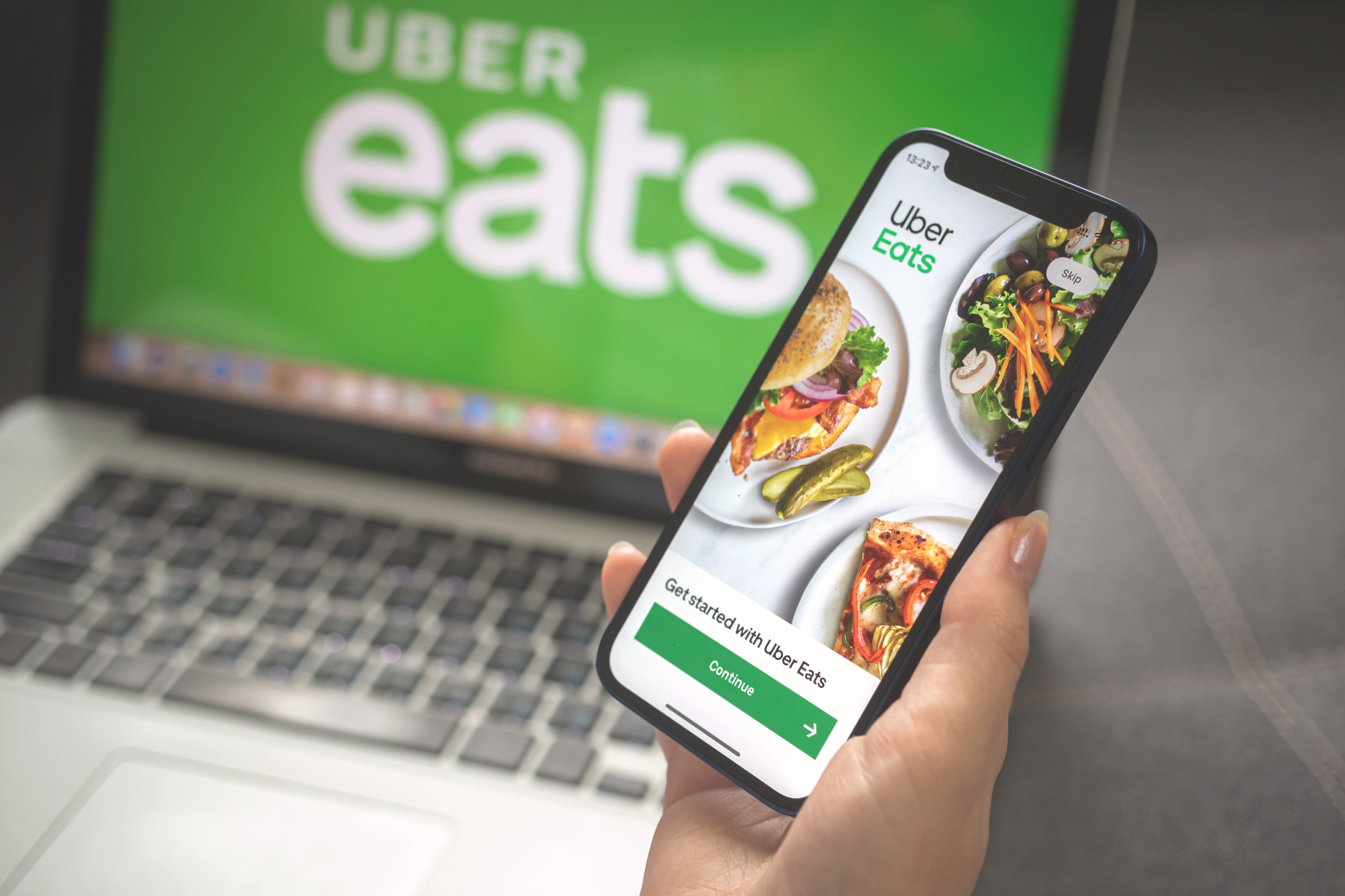Hand holding smartphone displaying Uber Eats app with food images and green Continue button, laptop in background